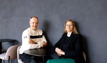 Capgeminin henkilöstöjohtaja Leena Kirjavainen ja luottamushenkilö Klaus West istuvat pienen mustan pöydän ääressä ja katsovat hymyillen kameraan.